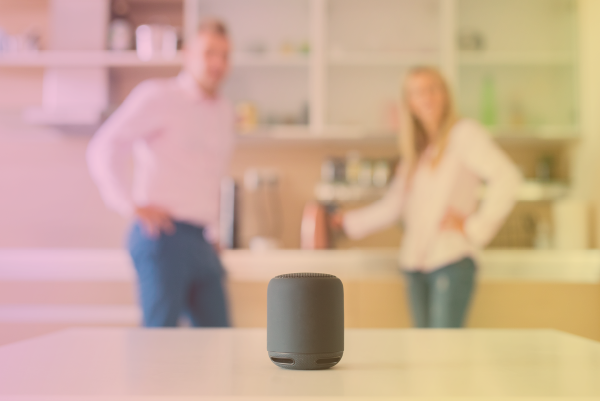 Man and woman in background talking to a smart speaker assistant