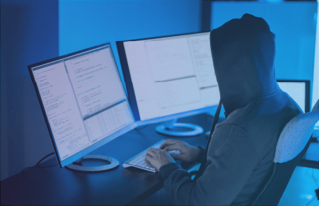Hooded figure sitting in front of computer with two monitors
